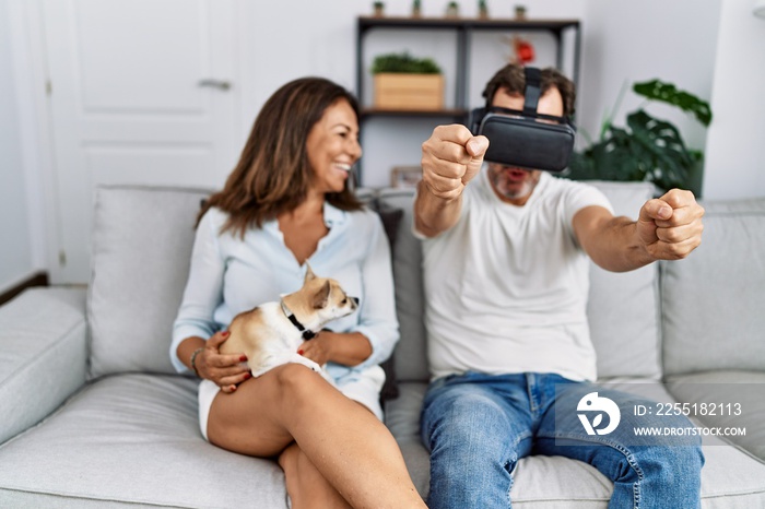 Middle age man and woman couple using vr goggles sitting on sofa with dog at home