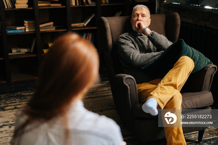 Thoughtful mature gray-haired man sits in front of unrecognizable young woman in cozy living room. Attractive red-haired young woman patient listening to older senior doctor explaining treatment.