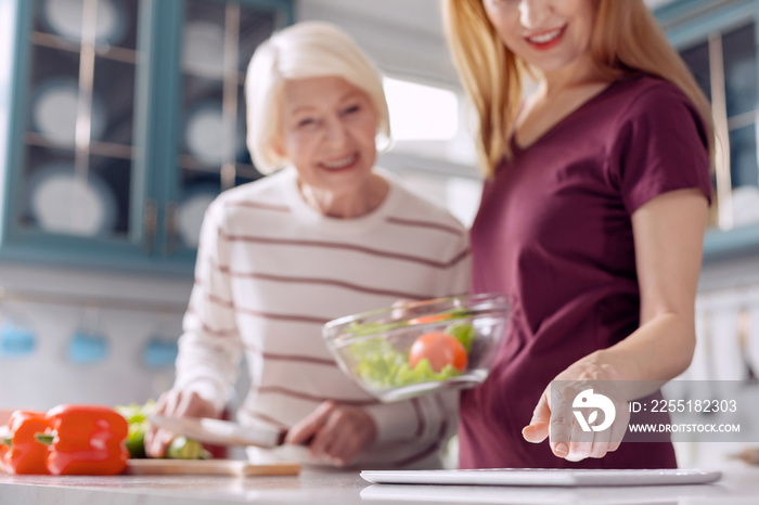 Helpful device. The focus being on the hand of a pleasant young woman checking a recipe on the tablet while making a salad with her elderly mother