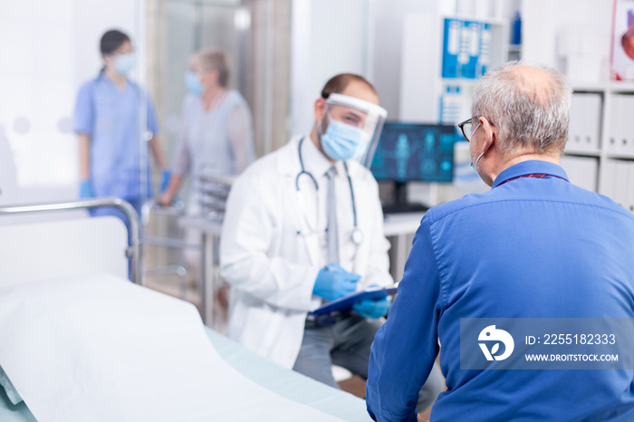 Old patient listening doctor diagnosis while wearing protective mask against coronavirus outbreak in hospital examination room. Modern private clinic . Practitioner physician appointment.