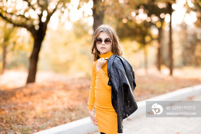 Cute stylish child girl 4-5 year old wear yellow knitted dress and sun glasses holding black leather jacket posing in park outdoor. Fall season. Childhood.