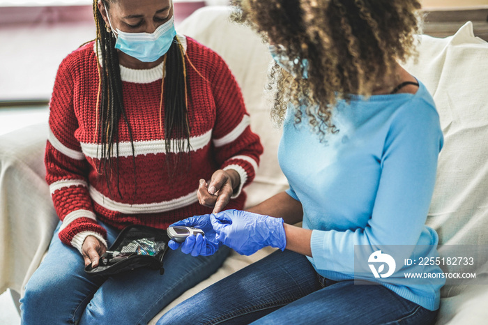 Young nurse woman and senior african patient waiting for the diabetes test result while wearing surgical face mask - Home isolated for coronavirus outbreak