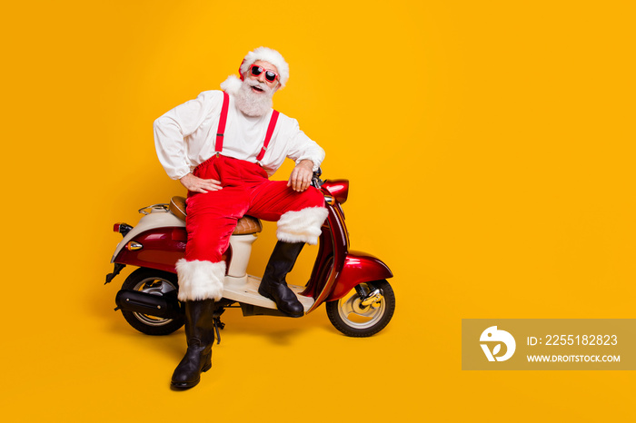 Full size photo of funny santa aged man in festive mood ready for x-mas theme party sitting on vintage bike wear sun specs pants cap shirt boots isolated yellow color background