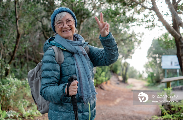 Smiling senior woman walking outdoors in a mountain path enjoying freedom and nature. Cheerful senior woman with backpack hiking in park making positive gesture with fingers