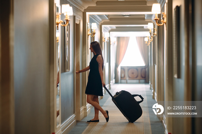 Young woman with handbag and suitcase in an elegant suit walks the hotel corridor to her room.