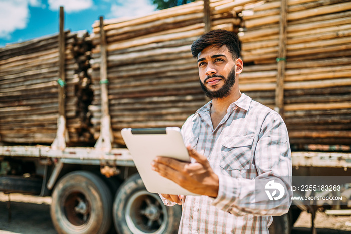 Portrait of Latin young man working with a digital tablet beside tree trunks on truck.