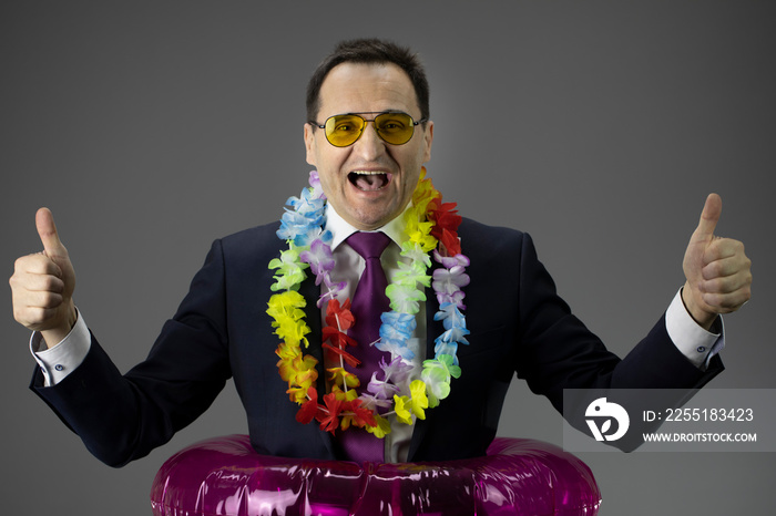 happy businessman dressed in a formal suit and lei Hawaiian welcome necklace in inflatable swim ring gesticulating in sunglasses isolated on gray background . holiday concept