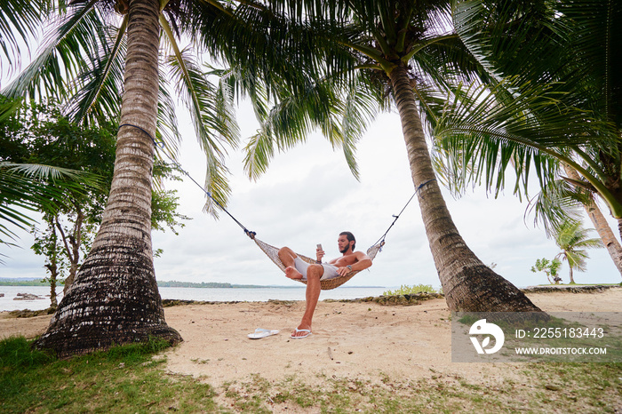 Tropical vacation. Young man relaxing at hammock on tropical beach.