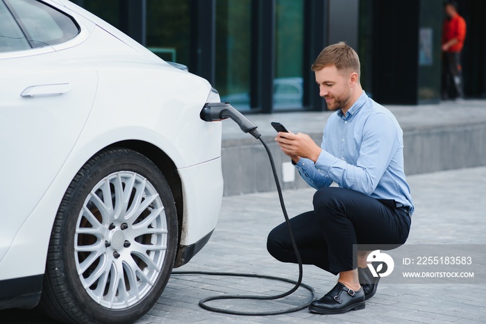 Man charging his electric car at charge station and using smartphone