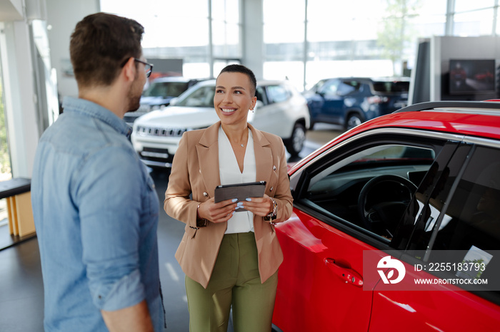 Handsome man with car dealer in auto show or salon. Saleswoman is holding a tablet in hands.