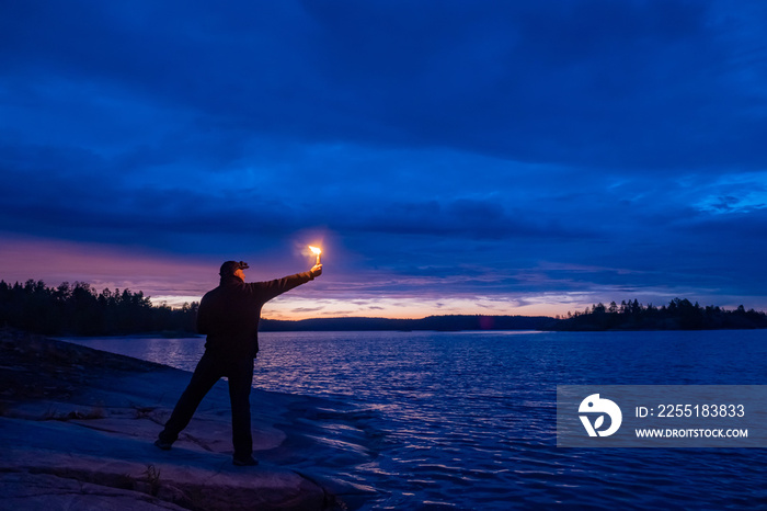 A man gives a signal with a smoke bomb. Smoke signals in the night. A man with a smoke torch by the water. Distress signal. The SOS signal. Call for help. Dangerous situation.