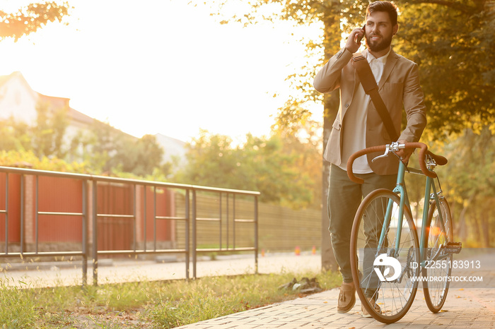 Young bearded businessman with bicycle talking by mobile phone on city street