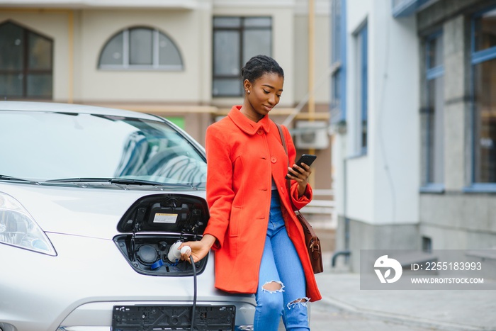 Charging electro car at the electric gas station. African American girl standing by the car.