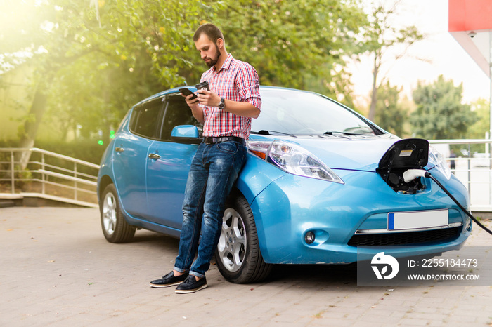 Young handsome man use phone and drink coffee while his electro car charging at station.