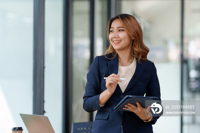 Attractive smiling Asian businesswoman standing holding tablet working and recording work details in office.