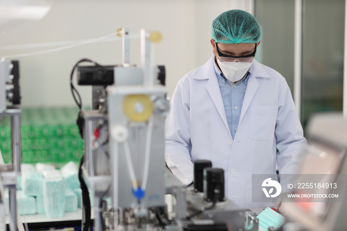 Worker in personal protective equipment or PPE inspecting quality of mask and medical face mask production line in factory, manufacturing industry and factory concept.