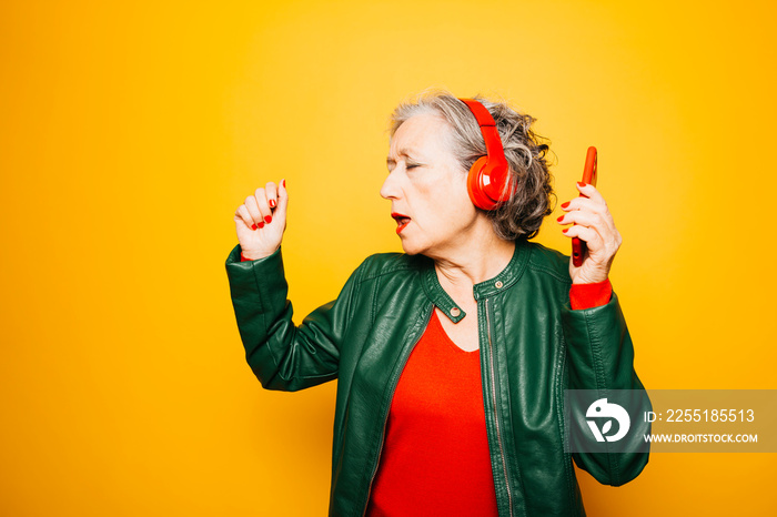 Senior woman with red headphones and red phone, dancing over a yellow background