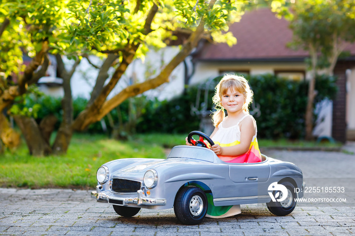 Little adorable toddler girl driving big vintage toy car and having fun with playing outdoors. Gorgeous happy healthy child enjoying warm summer day. Smiling stunning kid playing in domestic garden