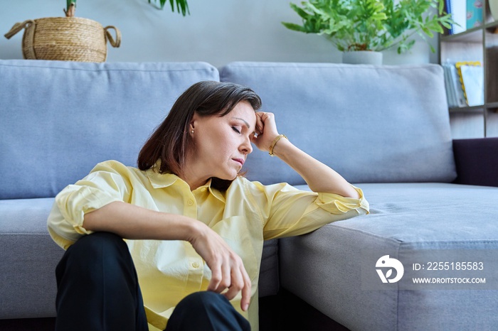 Sad upset middle-aged woman sitting on the floor near the sofa