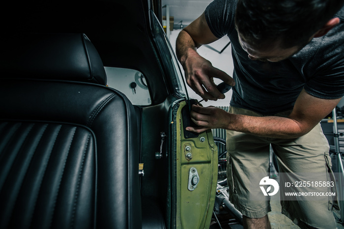 Young man removing interior trim panels from an old vintage car from the 60s or 70s in his home garage. Tools are seen around.