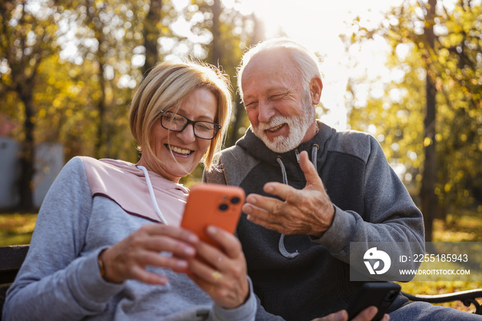 Cheerful seniors in sports clothing sitting on a park bench after exercise looking at smartphones, talking, and smiling. An active elderly couple in the park using a smartphone