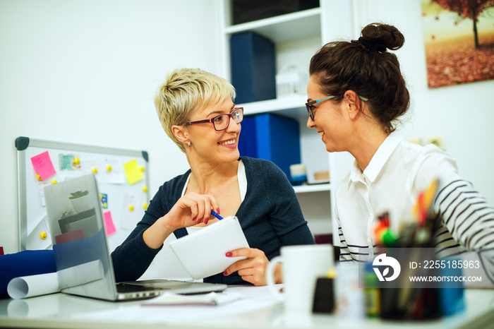 Two beautiful middle aged women colleague working together and looking each other while sitting at the desk in the office.