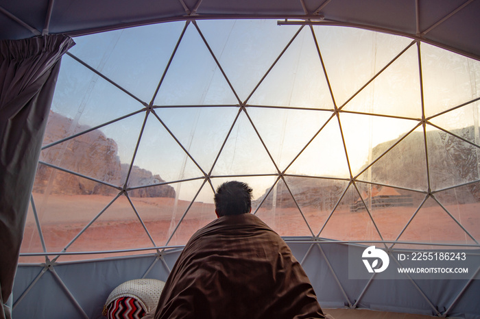 Tourist man staying in blanket in dome tent looking outside at Wadi Rum desert, famous natural attraction in Jordan. Travel Middle east concept