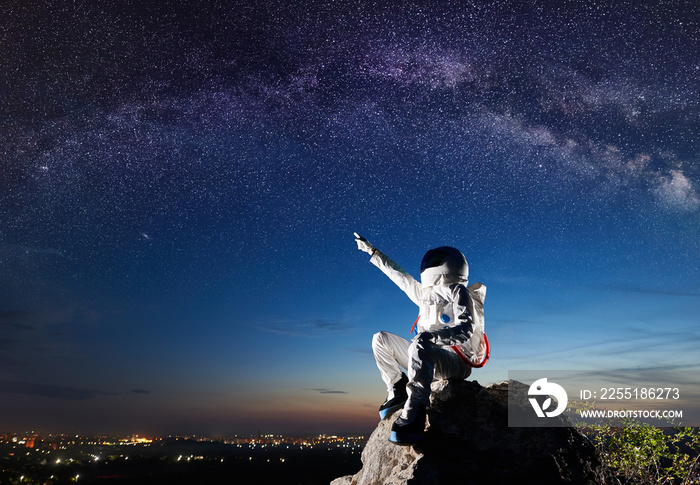 Side view of spaceman pointing at beautiful sky with stars while sitting on top of mountain. Mission specialist astronaut wearing white space suit with helmet. Concept of space travel and night sky.