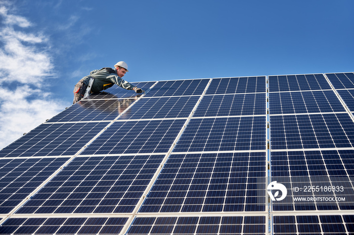 Horizontal snapshot of leaned workman in uniform and helmet, standing on ladder on the top of solar plant, installing solar modules on sunny day. Concept of alternative energy sources and innovations.