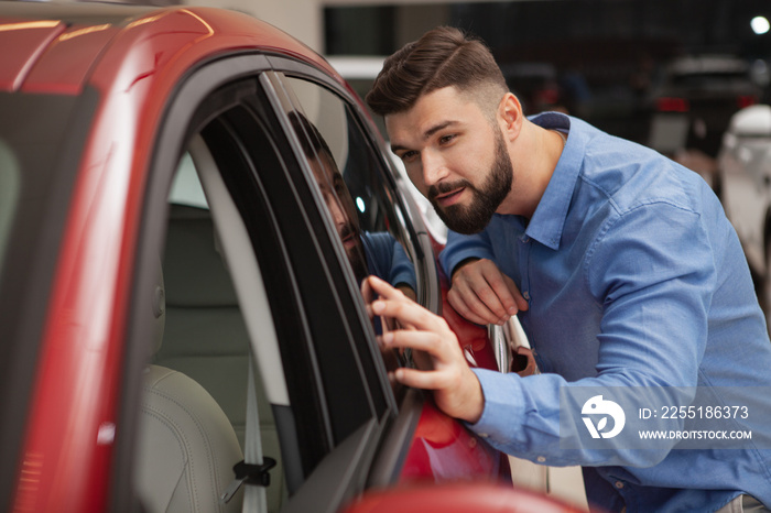 Handsome bearded young man examining new modern car at the dealership, copy space