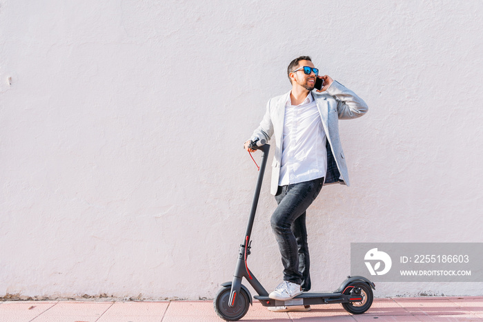 Latin adult man with sunglasses, well dressed and electric scooter talking on his mobile phone sitting on the street with a white wall background