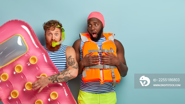 Photo of red haired shocked man wears snorkel mask, carries inflated mattress, shocked Afro American guy looks worried at friend, afraids of swimming, has no experience. Blank space on right