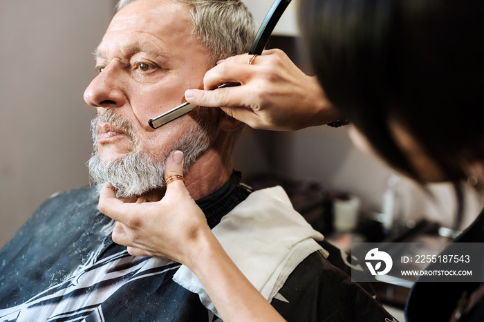 Professional barber girl shaves beard with a straight razor beard of mature man.