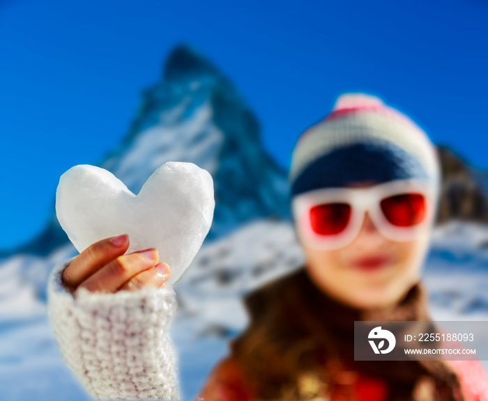 I love winter. Portrait of happy young girl holding in hand snow heart in winter time, ski slope in the background. Zermatt, Switzerland, Alps.