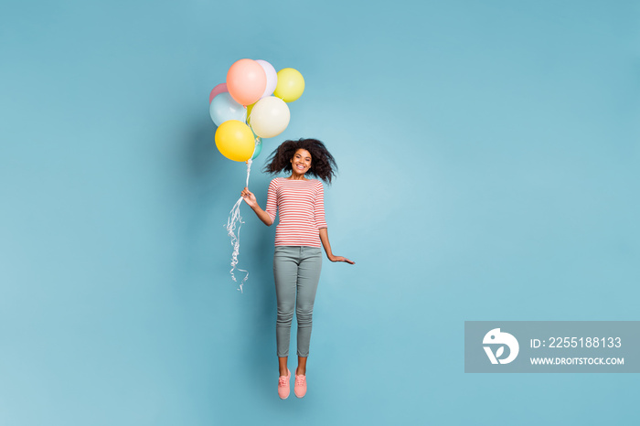 Photo of curly wavy cheerful trendy nice young girl wearing pants trousers striped t-shirt flying up holding baloons isolated over blue pastel color background