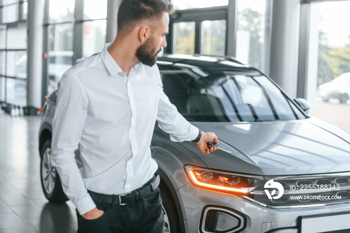 Using the keys remote controller. Young man in white clothes is in the car dealership