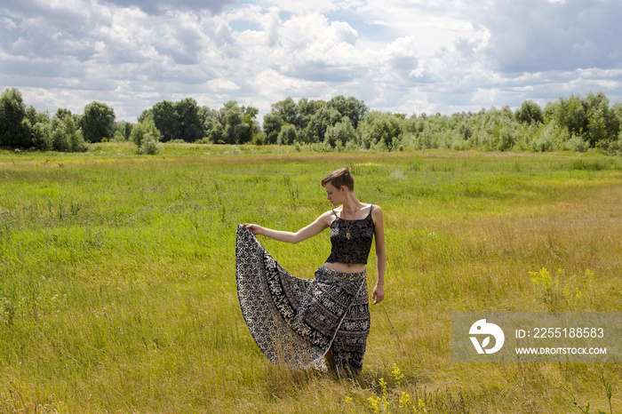 A girl in a long skirt with ethno patterns, raises her skirt edge with one hand, against the background of a field with flowers