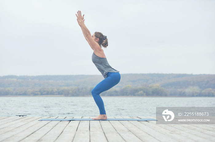 Beautiful young woman practices yoga asana Utkatasana - Chair pose on the wooden deck near the lake