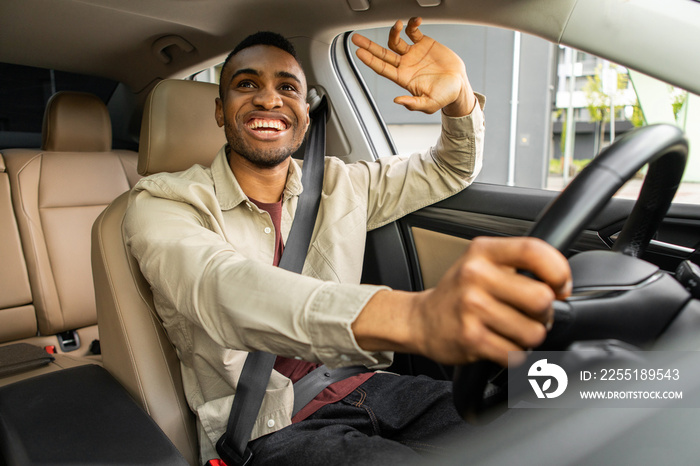 Young african american man smiling while driving a car, waving hello.