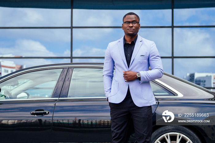 successful businessman handsome African American man in a stylish suit in a blue jacket standing in front of a cool new black car on the street