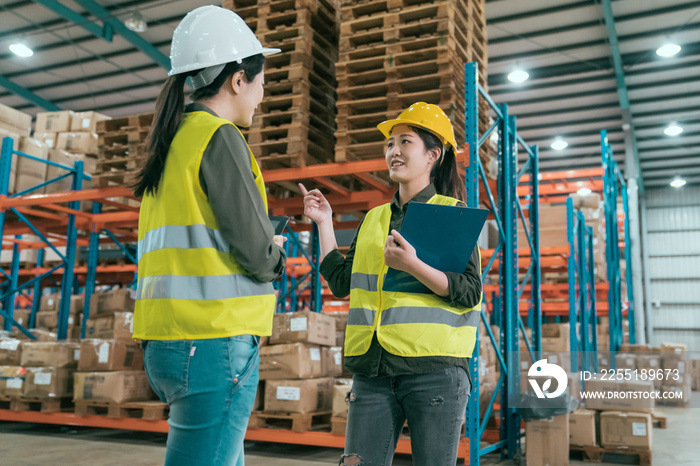 Happy female manager and worker standing together in warehouse office. two women staff in storehouse discussing cooperation. young girls employees wearing hard hats and safety vest in stockroom.