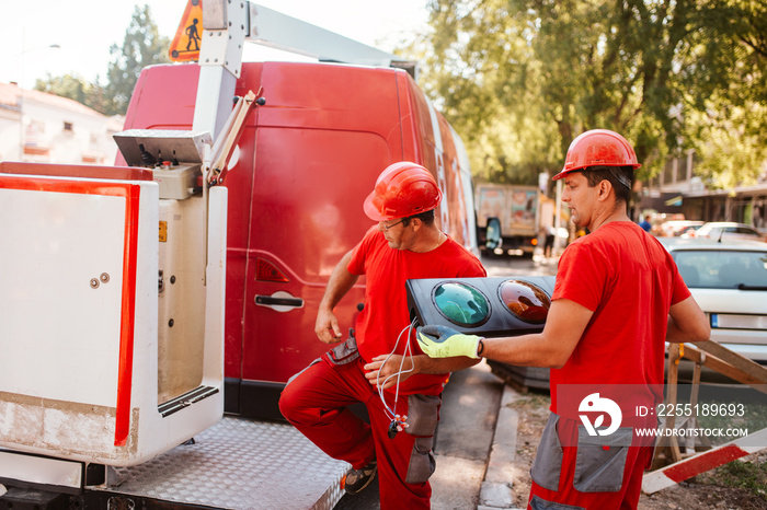 A middle-aged caucasian worker in a red uniform and helmet passes a traffic light to another worker. Setting up traffic lights on the street in progress