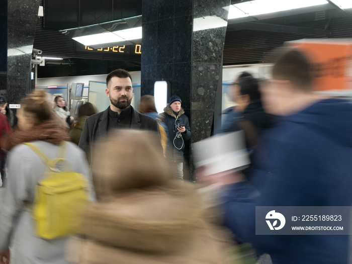 Handsome bearded man dressed in wool coat stands still in metro station within moving crowds of people