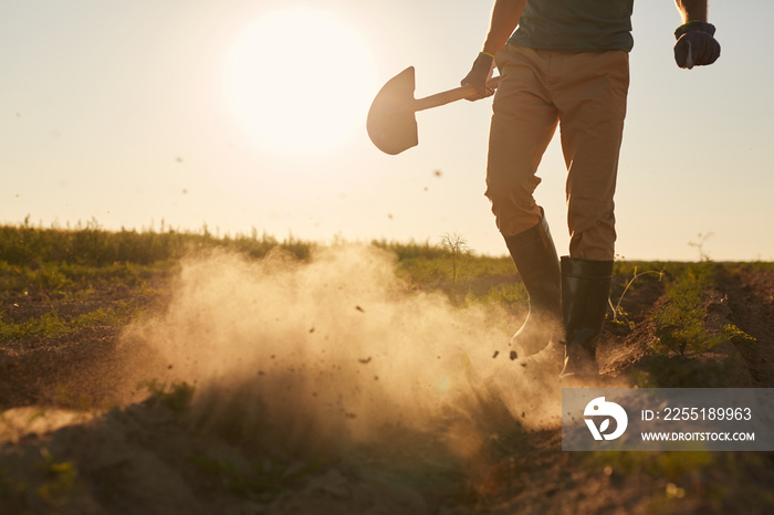 Low section portrait of unrecognizable male worker blowing clouds of dust from rubber boots and holding shovel while walking across plantation field in sunset light, copy space