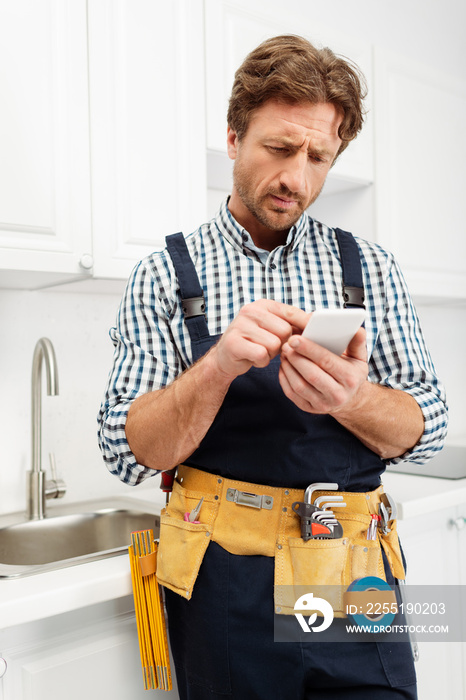 Selective focus of handsome plumber in tool belt and overalls using smartphone in kitchen