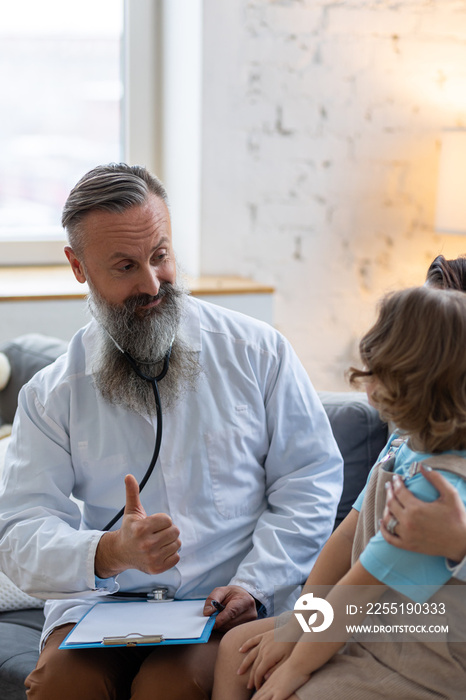 Kind male senior paediatrician doctor visiting his patient at home, examining little girl sitting on mother’s lap, writing prescription. Concept of kid’s health check. Sucessful recovery