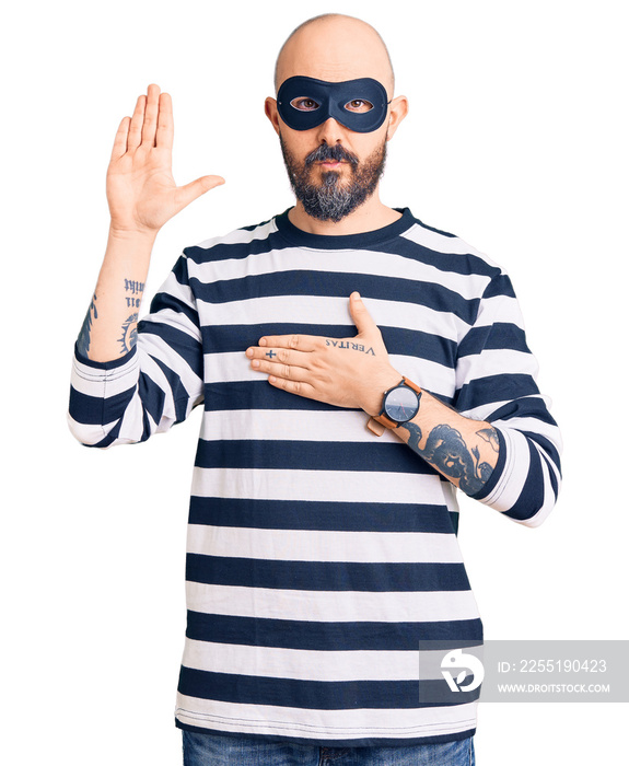 Young handsome man wearing burglar mask swearing with hand on chest and open palm, making a loyalty promise oath