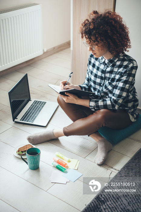 Upper view photo of a caucasian woman with curly hair and eyeglasses doing homework at home on the floor drinking a tea with sandwiches