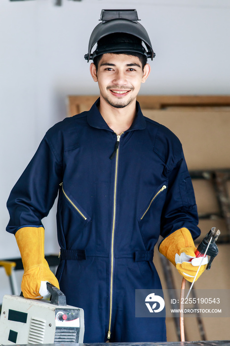 Happy young Asian male welder holds welding torch at workplace. Workshop and construction safety concept.