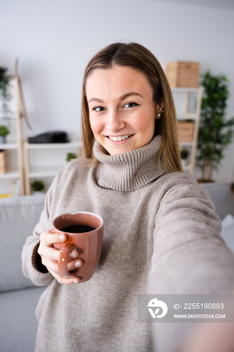 Beautiful blonde woman taking selfie at home while holding cup of coffee